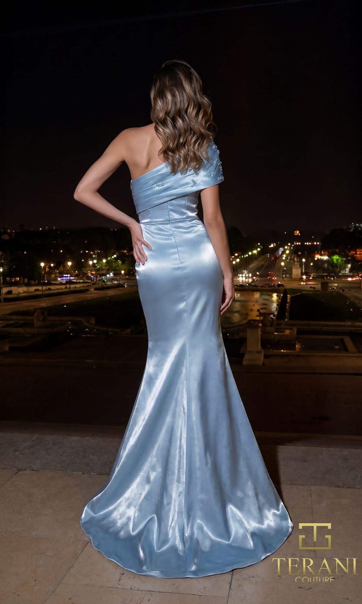 A woman stands outdoors at night in a shiny, light blue Long Formal Dress: Terani 261E6227, facing away from the camera with city lights twinkling in the background.